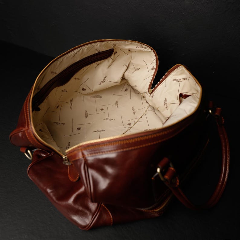 K&ouml;nnte beinhalten: Eine braune Leder-Reisetasche mit Rei&szlig;verschluss oben und zwei Griffen. Die Tasche ist mit einem cremefarbenen Stoff gef&uuml;ttert, auf dem immer wieder die Worte "Made in Italy" stehen.
