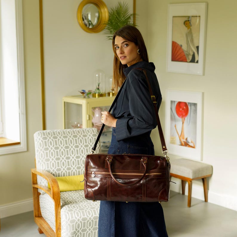 K&ouml;nnte beinhalten: Eine braune Reisetasche aus Leder mit einem langen Trageriemen. Die Tasche hat zwei obere Griffe und einen Rei&szlig;verschluss.
