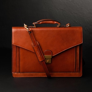 May include: A brown leather briefcase with a gold clasp and a long strap. The briefcase has a flap that closes with a buckle.