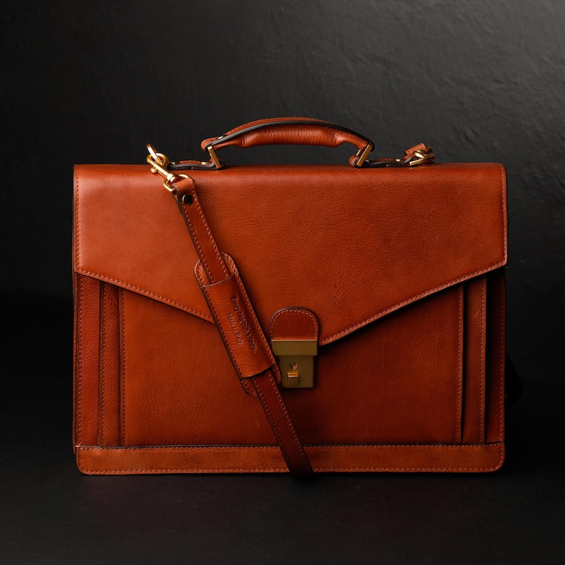 May include: A brown leather briefcase with a gold clasp and a long strap. The briefcase has a flap that closes with a buckle.