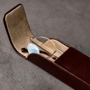 May include: A dark brown rectangular glasses case with a hinged lid standing open to reveal a beige interior lining. Inside the case rests a pair of round-framed glasses with thin metallic rims and dark brown temple arms visible.