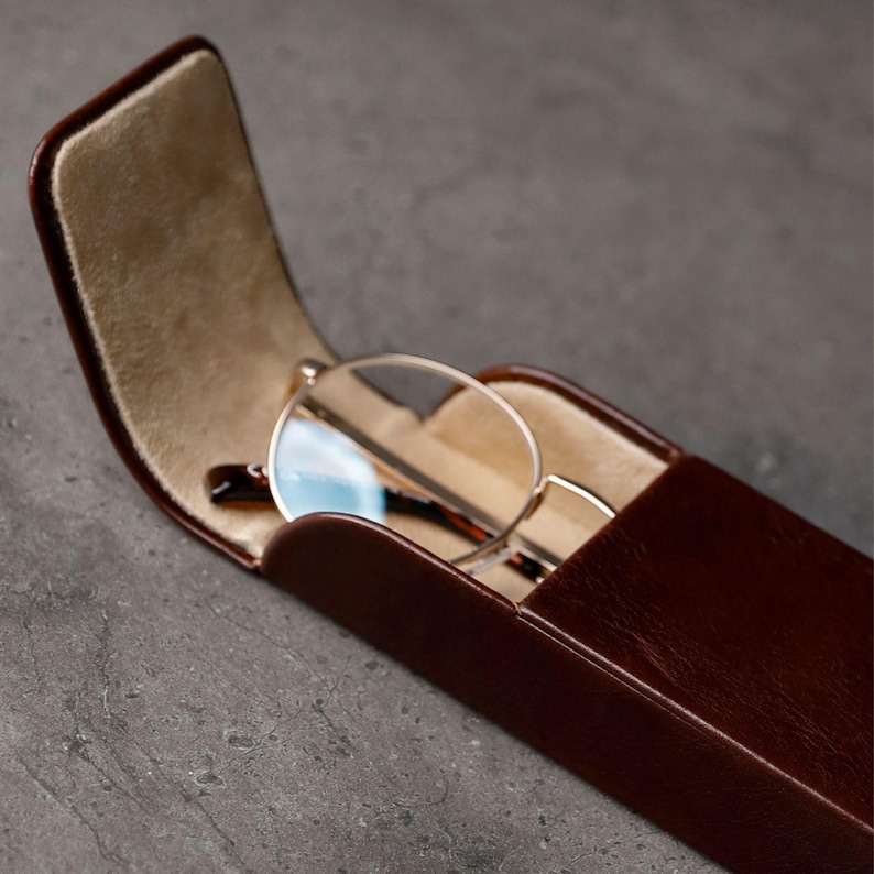 May include: A dark brown rectangular glasses case with a hinged lid standing open to reveal a beige interior lining. Inside the case rests a pair of round-framed glasses with thin metallic rims and dark brown temple arms visible.