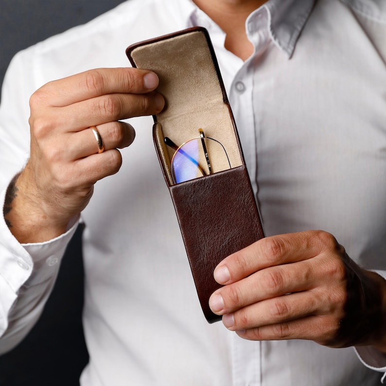 May include: A dark brown, vertical, rectangular glasses case with a hinged lid, featuring a light beige interior lining. Inside the case, a pair of gold-rimmed glasses with blue-tinted lenses is visible, partially tucked into the protective sleeve.