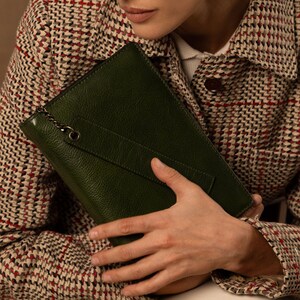 May include: A dark green rectangular clutch bag with a textured leather-like surface, featuring a diagonal strap across the front and a small metallic chain attachment connected to a circular metal ring on the upper left corner of the bag.