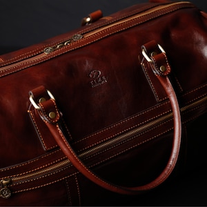 K&ouml;nnte beinhalten: Eine braune Reisetasche aus Leder mit goldenen Rei&szlig;verschl&uuml;ssen und Beschl&auml;gen. Die Tasche hat einen oberen Griff und ein Markenlogo mit der Aufschrift "T.I.L.A.  Leatherware".