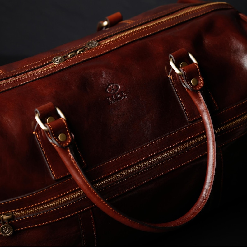 K&ouml;nnte beinhalten: Eine braune Reisetasche aus Leder mit goldenen Rei&szlig;verschl&uuml;ssen und Beschl&auml;gen. Die Tasche hat einen oberen Griff und ein Markenlogo mit der Aufschrift "T.I.L.A.  Leatherware".