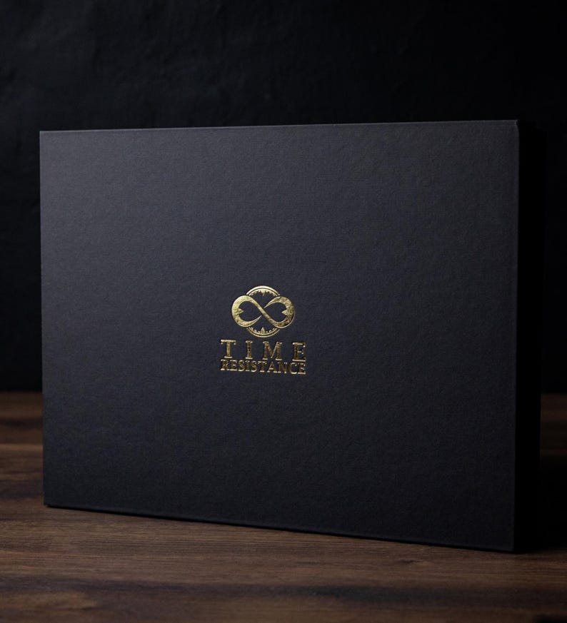 Puede incluir: Una caja rectangular negra con un logotipo dorado que dice "TIME RESISTANCE". El logotipo presenta un dise&ntilde;o de s&iacute;mbolo de infinito. La caja est&aacute; sobre una superficie de madera.