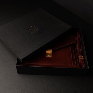 May include: A burgundy leather portfolio with a gold clasp and strap, inside a black box with the logo "The  Fox & The  Hare".