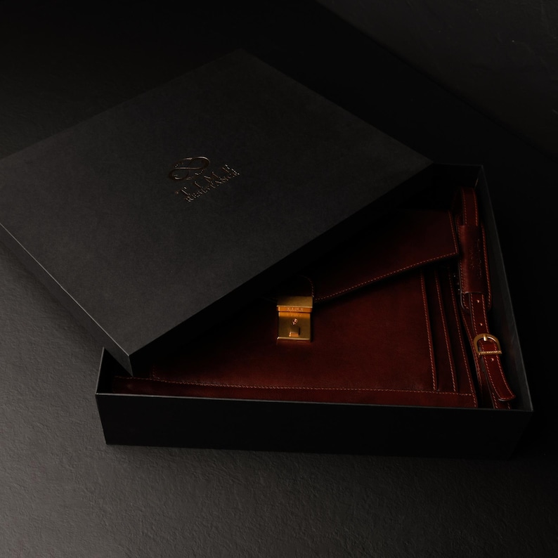 May include: A burgundy leather portfolio with a gold clasp and strap, inside a black box with the logo "The  Fox & The  Hare".