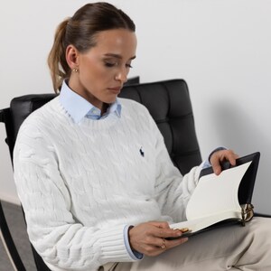May include: A white cable-knit sweater with a small navy blue embroidered logo on the chest, worn over a light blue collared shirt. The sweater features a crew neckline and long sleeves with ribbed cuffs, paired with light-colored trousers.