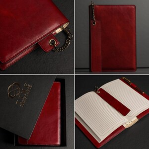 May include: A deep red leather notebook cover featuring a metallic chain attachment and a matching leather bookmark strap. The notebook includes lined cream-colored pages and is presented in a black box with gold embossed branding on the lid.