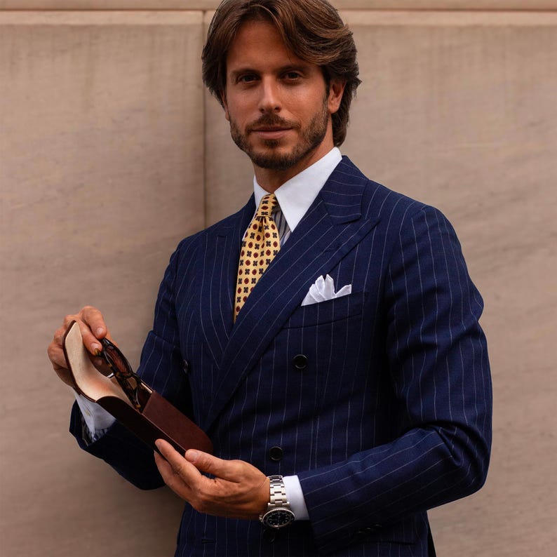 May include: A navy blue double-breasted pinstripe blazer with a white collared shirt, a yellow patterned tie, and a white pocket square. A silver wristwatch is worn on the wrist, and a brown leather glasses case is held in the hand.