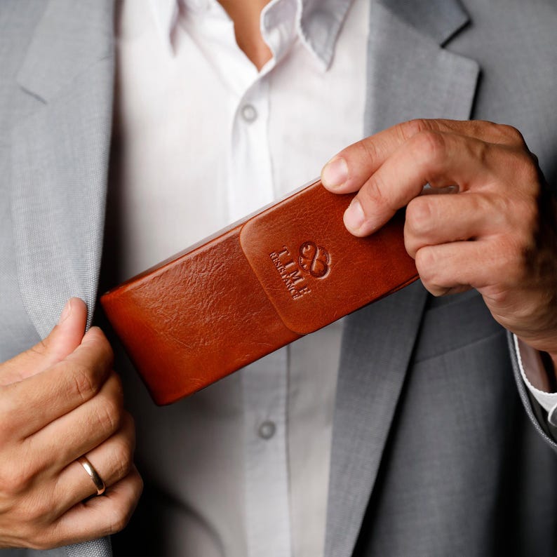 May include: A rectangular brown leather glasses case with a smooth texture and a flap closure. The front of the case features an embossed logo with the text TIME RESISTANCE and a stylized infinity symbol centered on the flap surface.