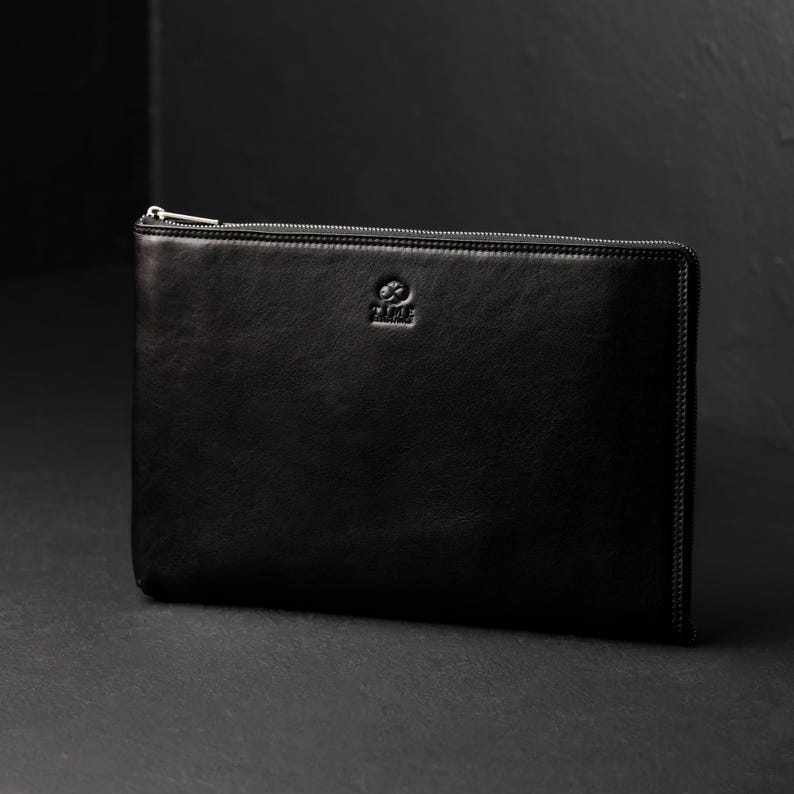 Puede incluir: Un malet&iacute;n de documentos de cuero negro con cierre de cremallera plateado. El malet&iacute;n rectangular tiene una textura suave y un peque&ntilde;o logotipo en relieve en la parte delantera. Dise&ntilde;ado para guardar documentos y otros elementos esenciales.