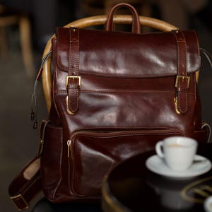 K&ouml;nnte beinhalten: Ein dunkelbrauner Lederrucksack mit einer oberen Klappe, zwei vertikalen Riemen, goldfarbenen Metallschnallen und einer vorderen Rei&szlig;verschlusstasche. Der Rucksack verf&uuml;gt &uuml;ber einen Tragegriff, verstellbare Schultergurte und Seitentaschen mit Druckkn&ouml;pfen.