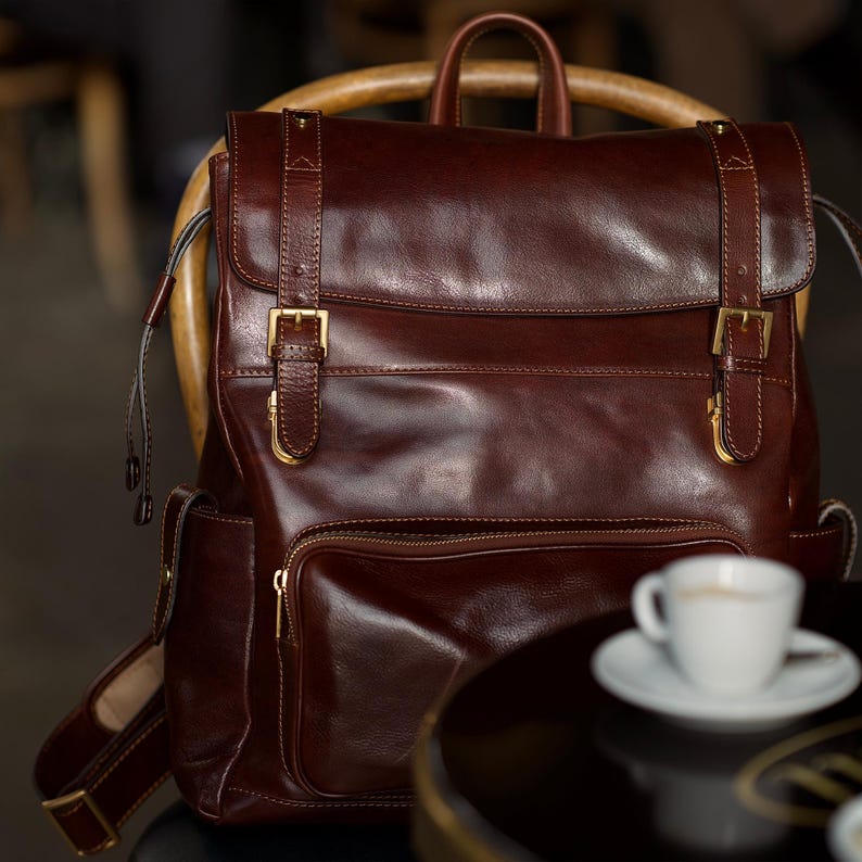 K&ouml;nnte beinhalten: Ein dunkelbrauner Lederrucksack mit einer oberen Klappe, zwei vertikalen Riemen, goldfarbenen Metallschnallen und einer vorderen Rei&szlig;verschlusstasche. Der Rucksack verf&uuml;gt &uuml;ber einen Tragegriff, verstellbare Schultergurte und Seitentaschen mit Druckkn&ouml;pfen.