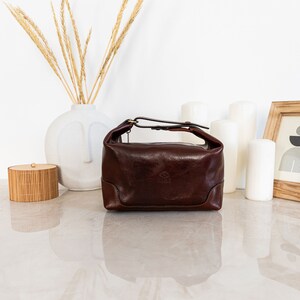 May include: A brown leather travel toiletry bag with a top handle and a buckle closure. The bag has a small logo on the front.