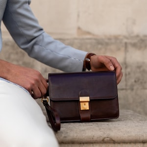 May include: A dark brown leather clutch bag featuring a rectangular silhouette, a fold-over flap, and a polished gold-tone metal push-lock closure. The bag includes a matching leather wrist strap attached to a gold-tone metal ring on the side.