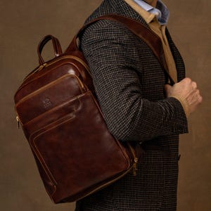 May include: A dark brown leather backpack featuring a structured rectangular shape, a top handle, and gold-toned metal zips. The bag has a front zipped pocket with visible stitching details and a small embossed logo on the upper front section.