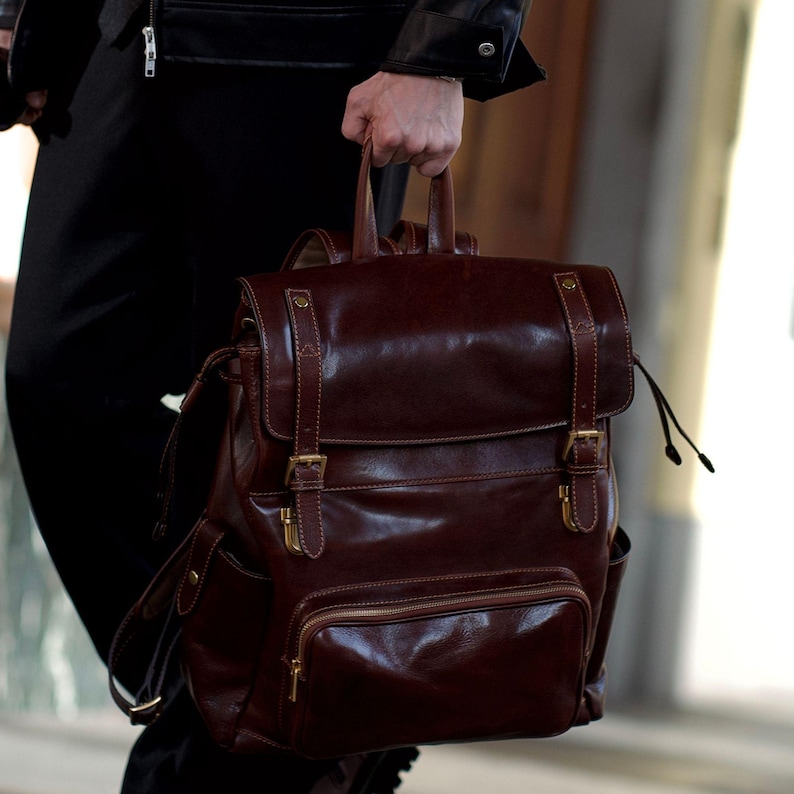 K&ouml;nnte beinhalten: Ein dunkelbrauner Lederrucksack mit einem &Uuml;berschlag, zwei vertikalen Riemen und goldfarbenen Metallschnallen. Die Tasche verf&uuml;gt &uuml;ber eine vordere Rei&szlig;verschlusstasche, Seitentaschen und einen Tragegriff mit einer polierten Oberfl&auml;che.
