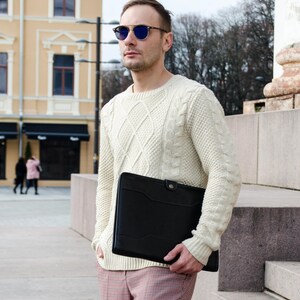 May include: A man wearing a white cable knit sweater and pink and black plaid pants holds a black leather portfolio. He is wearing blue-tinted sunglasses.