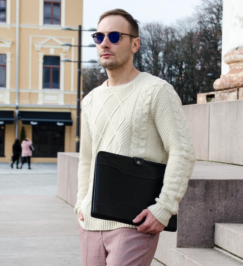 May include: A man wearing a white cable knit sweater and pink and black plaid pants holds a black leather portfolio. He is wearing blue-tinted sunglasses.