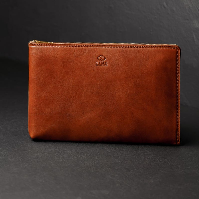 Puede incluir: Un bolso de mano rectangular de cuero marr&oacute;n con cierre de cremallera dorada. El bolso tiene una textura suave y un peque&ntilde;o logotipo en la parte superior central. El cuero es de color marr&oacute;n medio c&aacute;lido.