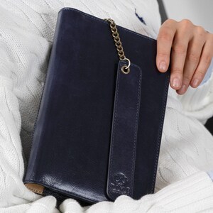 May include: A dark navy blue leather notebook cover featuring a vertical strap secured by a brass-toned metal chain. The cover displays an embossed infinity symbol logo above the word TIME at the bottom right corner, with visible stitching.