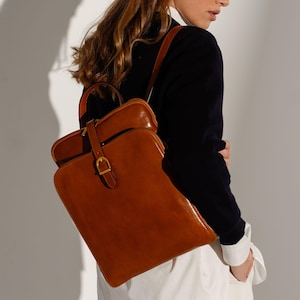 Pode incluir: Uma mochila de couro cor de conhaque com uma silhueta retangular fina de cantos arredondados, uma alça superior e uma aba frontal presa por uma tira de couro vertical com uma fivela de metal dourado. A bolsa tem um acabamento liso.