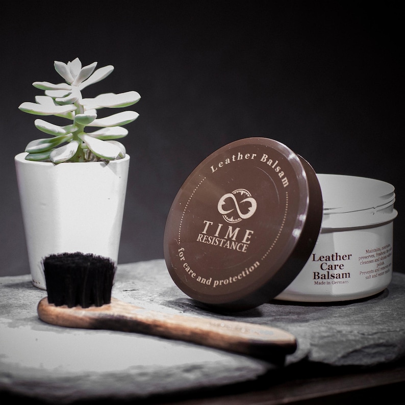 May include: A white jar of Time Resistance Leather Care Balsam with a brown lid featuring an infinity logo, accompanied by a wooden brush with black bristles. The text on the lid reads Leather Balsam for care and protection, Time Resistance.