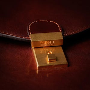 May include: Close-up of a brown leather briefcase with a gold metal clasp. The clasp has the text "TIMBER HERITAGE" engraved on it.