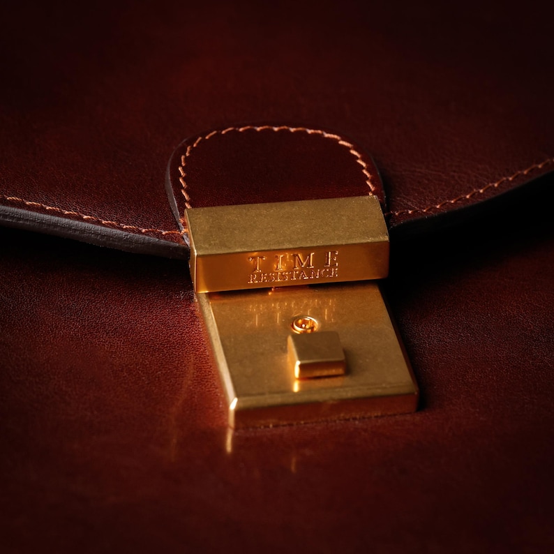 May include: Close-up of a brown leather briefcase with a gold metal clasp. The clasp has the text "TIMBER HERITAGE" engraved on it.