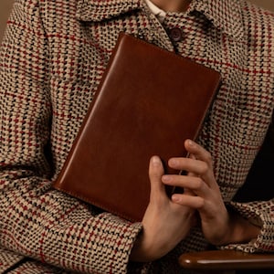 May include: A rectangular brown leather notebook with a smooth finish and a zipper closure along the top edge. The notebook is held against a textured beige, red, and black houndstooth patterned jacket with a visible dark brown button.