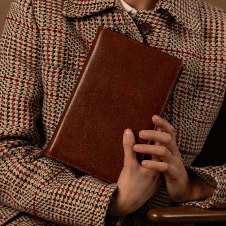 May include: A rectangular brown leather notebook with a smooth finish and a zipper closure along the top edge. The notebook is held against a textured beige, red, and black houndstooth patterned jacket with a visible dark brown button.