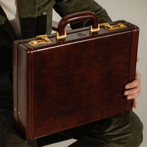May include: A dark brown leather briefcase featuring a structured rectangular shape, a sturdy top handle, and two gold-toned combination locks. The exterior surface has a smooth, polished finish with visible stitching along the edges.