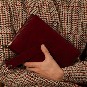 May include: A deep burgundy suede clutch bag featuring a rectangular silhouette, a matching wrist strap, and a thin metallic chain attachment. The bag is held against a beige, brown, and red houndstooth patterned fabric garment with a visible button.