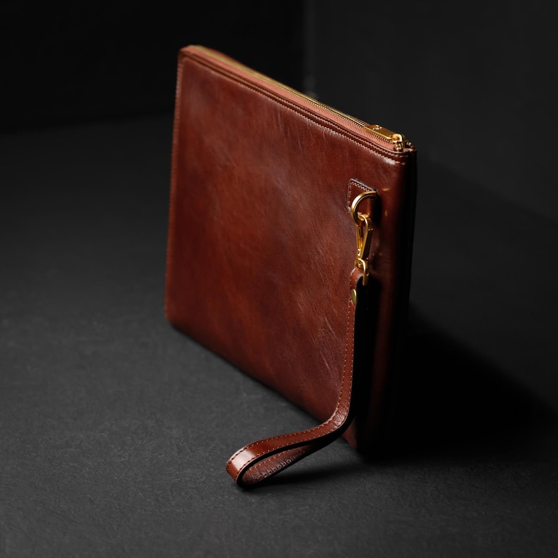 Puede incluir: Un bolso de mano de cuero marr&oacute;n con cremallera dorada y correa de mu&ntilde;eca. La bolsa rectangular est&aacute; hecha de cuero liso y pulido. La correa de mu&ntilde;eca est&aacute; sujeta con un cierre dorado.