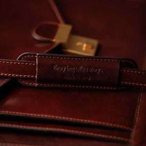 May include: Close-up of a brown leather bag with a gold clasp. The bag has a leather strap with the text "Every bag tells a story... Made in Italy" embossed on it.