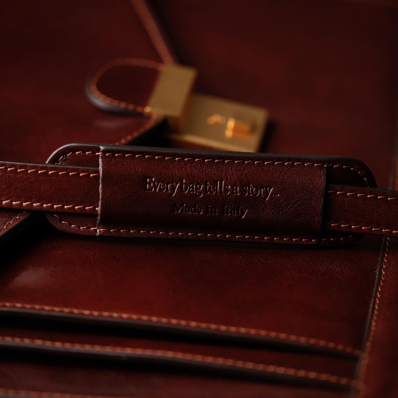 May include: Close-up of a brown leather bag with a gold clasp. The bag has a leather strap with the text "Every bag tells a story... Made in Italy" embossed on it.