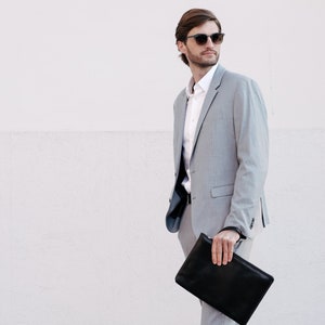 Puede incluir: Un traje gris claro con camisa blanca y gafas de sol negras. La persona sostiene un bolso de mano de cuero negro. El fondo es una pared blanca. La chaqueta del traje tiene cierre de dos botones.