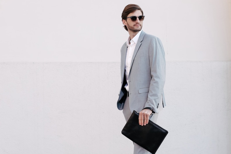 Puede incluir: Un traje gris claro con camisa blanca y gafas de sol negras. La persona sostiene un bolso de mano de cuero negro. El fondo es una pared blanca. La chaqueta del traje tiene cierre de dos botones.