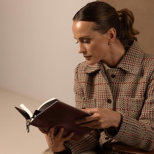 May include: A brown leather-bound book with a metal chain bookmark, held by a person wearing a beige, brown, and red plaid jacket with a collared neckline, button-down front, and visible chest pockets, featuring a textured, woven fabric pattern.