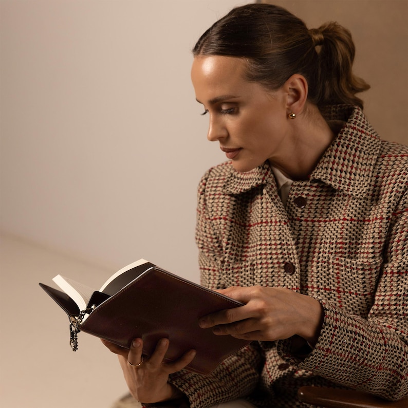 May include: A brown leather-bound book with a metal chain bookmark, held by a person wearing a beige, brown, and red plaid jacket with a collared neckline, button-down front, and visible chest pockets, featuring a textured, woven fabric pattern.