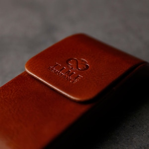 May include: A brown leather case with a smooth, polished finish and a rounded flap closure. The surface features a debossed logo with an infinity symbol above the text TIME RESISTANCE, clearly visible on the front flap of the leather accessory.
