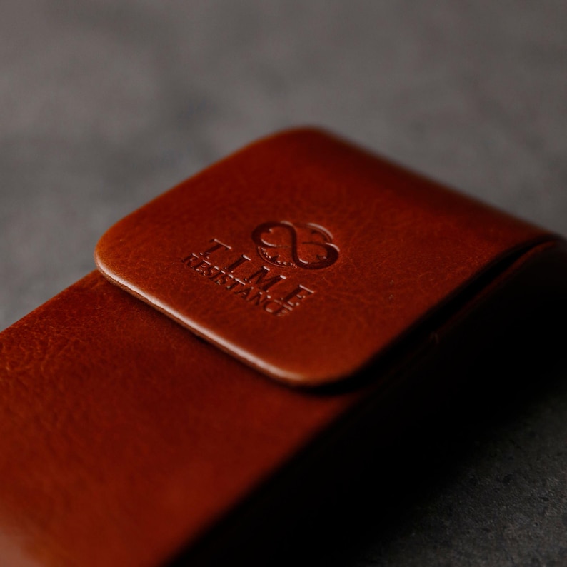 May include: A brown leather case with a smooth, polished finish and a rounded flap closure. The surface features a debossed logo with an infinity symbol above the text TIME RESISTANCE, clearly visible on the front flap of the leather accessory.