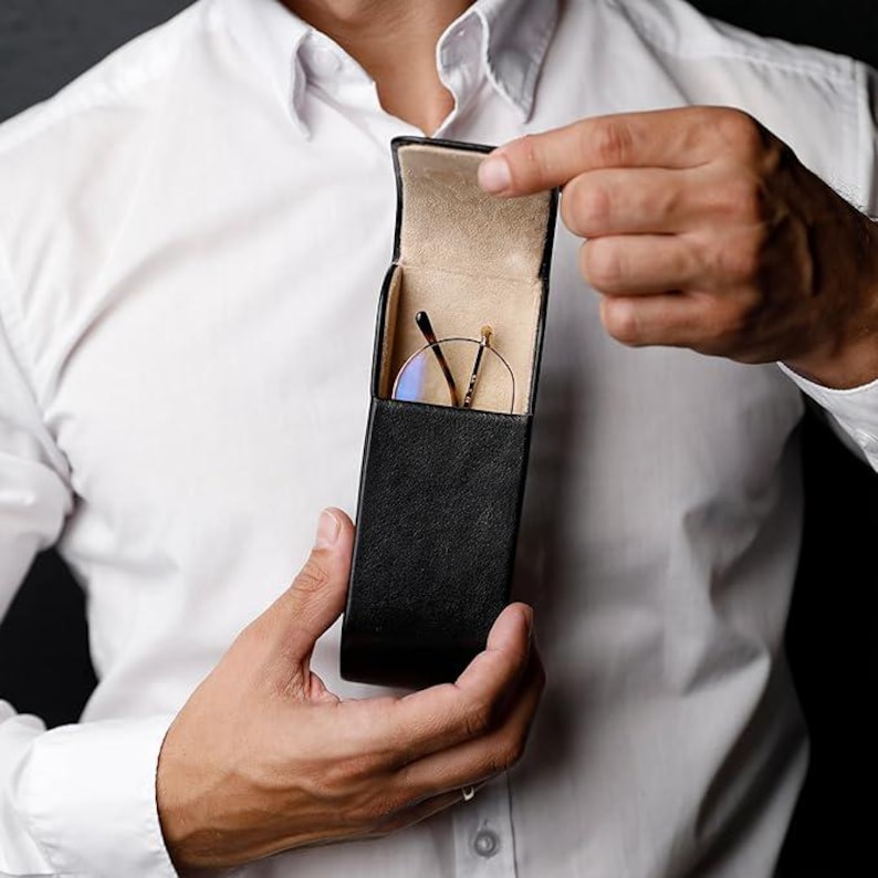 May include: A black leather-textured glasses case with a vertical rectangular shape and a hinged lid. The interior is lined with a light beige material, and a pair of thin-rimmed glasses with gold-toned arms is visible inside the open case.