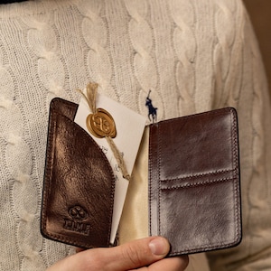 May include: A dark brown leather wallet with a glossy finish, featuring multiple card slots and a pocket containing a white card with a gold wax seal. The wallet is embossed with a logo and the text TIME, showing visible stitching along the edges.