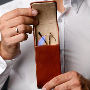 Könnte beinhalten: Ein vertikales Brillenetui aus braunem Leder mit einem aufgeklappten Deckel, das eine Brille mit rundem Rahmen, dünnen goldfarbenen Metallbügeln und blau getönten Gläsern im beigefarbenen, mit Filz ausgekleideten Innenfach zeigt.