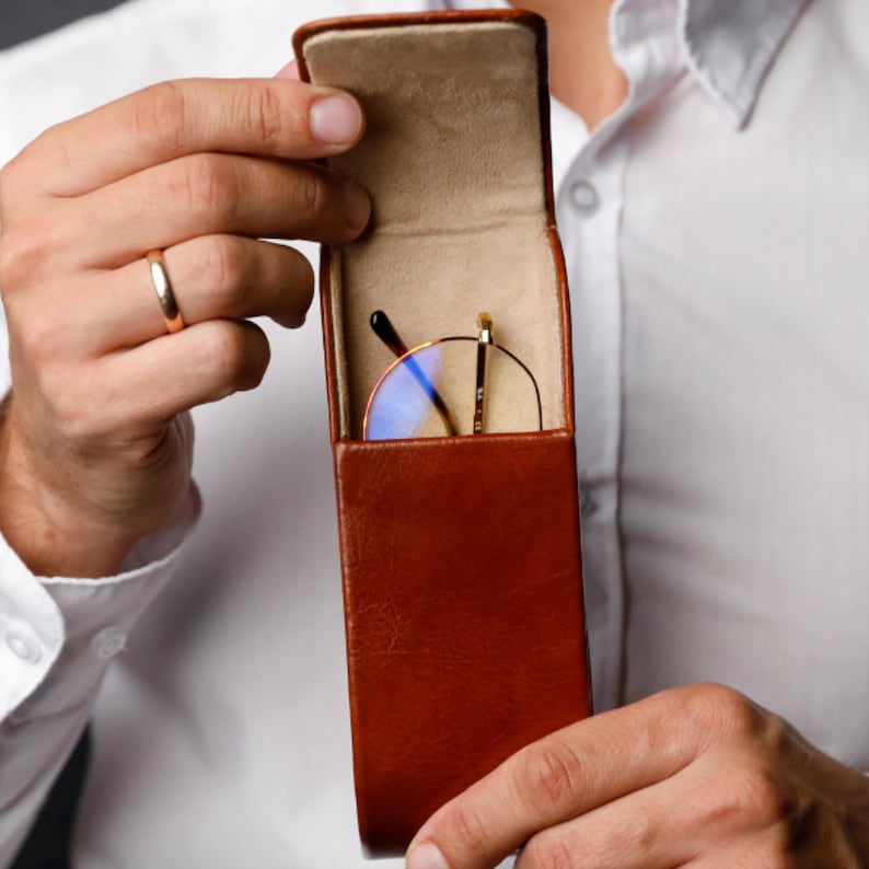 May include: A vertical brown leather glasses case with a hinged lid held open to reveal a pair of round-framed eyeglasses with thin gold metal temples and blue-tinted lenses resting inside the beige felt-lined interior compartment.