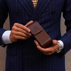 May include: A dark brown leather case with a flap closure and a debossed logo on the front. The case is held in front of a navy blue pinstriped double-breasted blazer with a white dress shirt and a patterned yellow tie visible at the collar.
